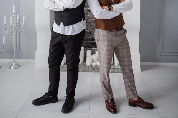 Two men in formal vests and trousers pose stylishly with arms crossed.