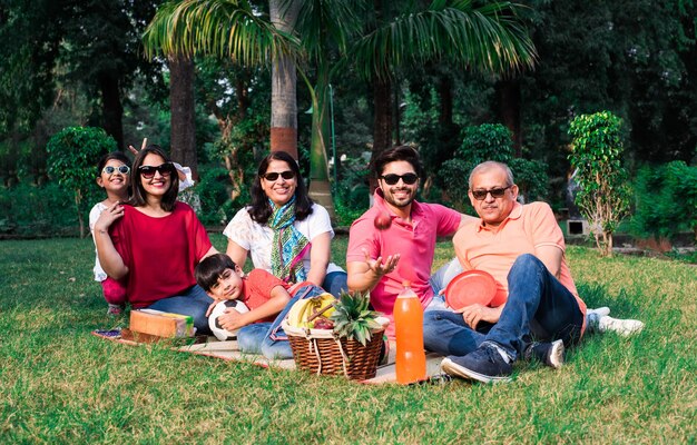 A multigenerational family sitting on a picnic mat in a park with food and drinks, smiling at the camera.