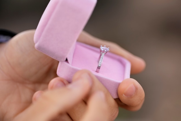 A hand holding an open pink ring box with a diamond engagement ring inside.