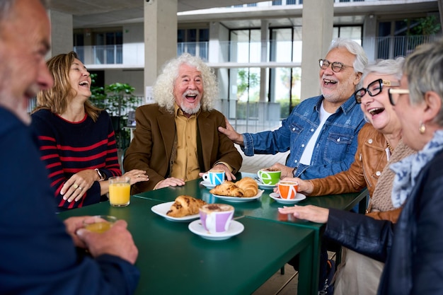 Group of smiling seniors laughing together at a café table, sharing coffee and pastries in a bright outdoor setting, joy.