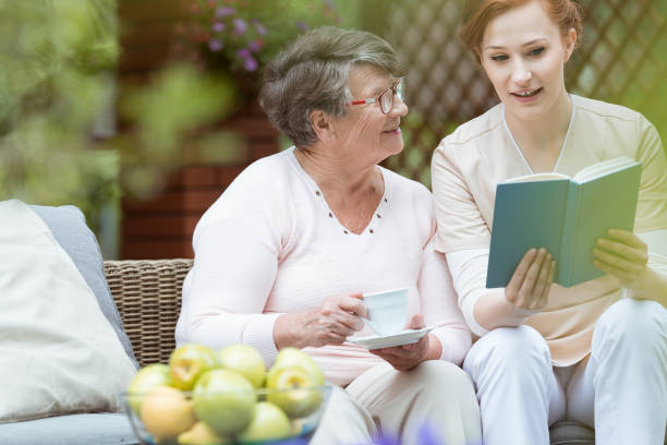 Caregiver reading a book with an elderly woman on a garden sofa, sharing tea, smiles, and a supportive moment outdoors.