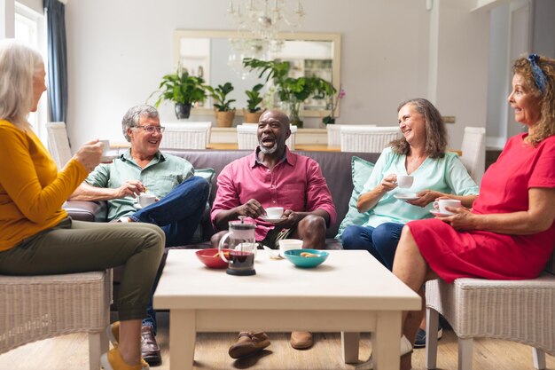 Group of seniors chatting and laughing over tea in a bright living room, enjoying friendship, comfort, and relaxed social time.