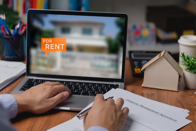 Hands signing rental agreement at desk with laptop showing For Rent home, small house model and coffee.
