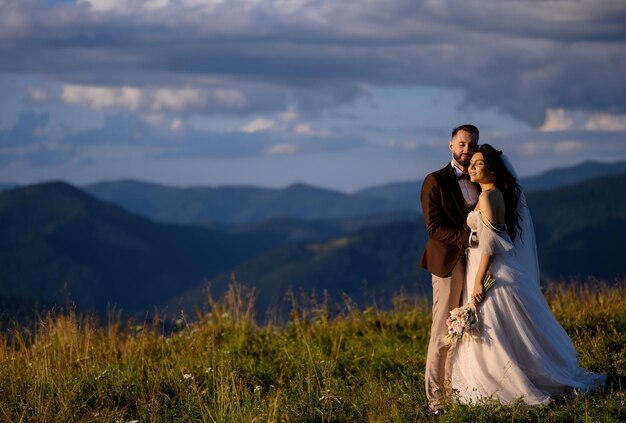 Romantic outdoor wedding portrait of newlyweds against dramatic mountains and evening sky.