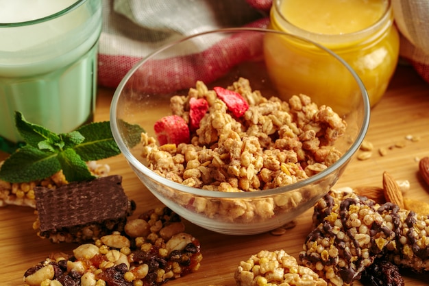 A bowl of crunchy granola with dried strawberries on a wooden table, surrounded by cereal bars, milk, and honey.