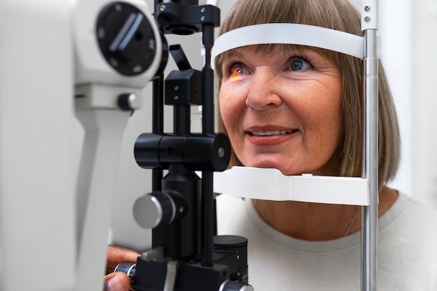 A woman is undergoing an eye exam using a slit lamp machine, smiling slightly as a bright light checks her vision.